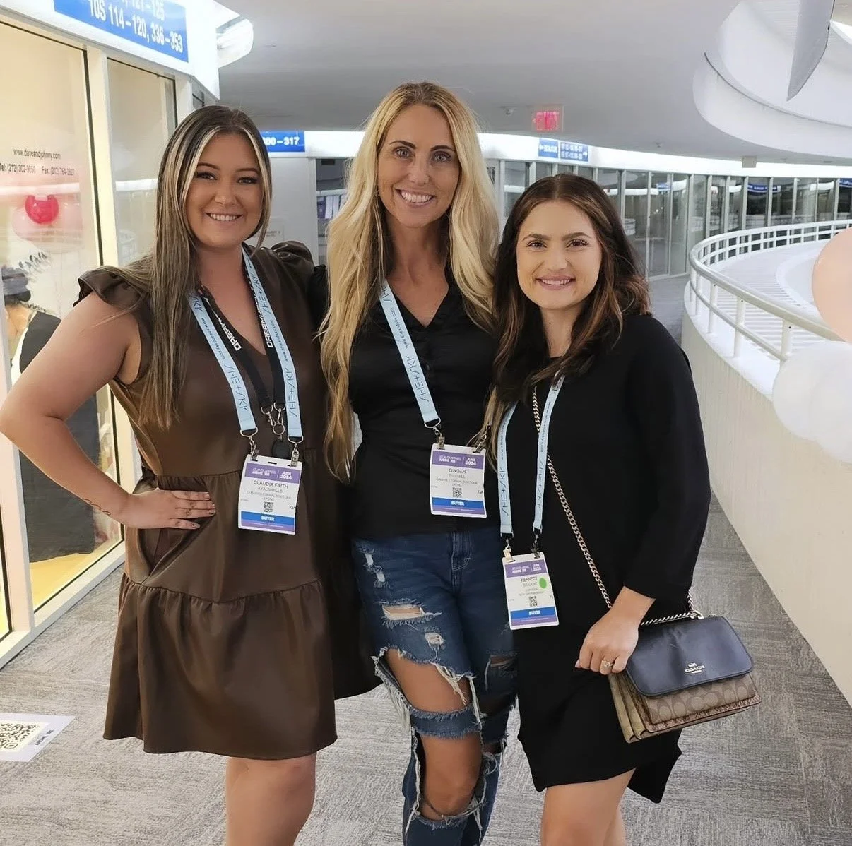Three women standing together inside a building, smiling. They are wearing conference badges on lanyards around their necks and casual clothing.