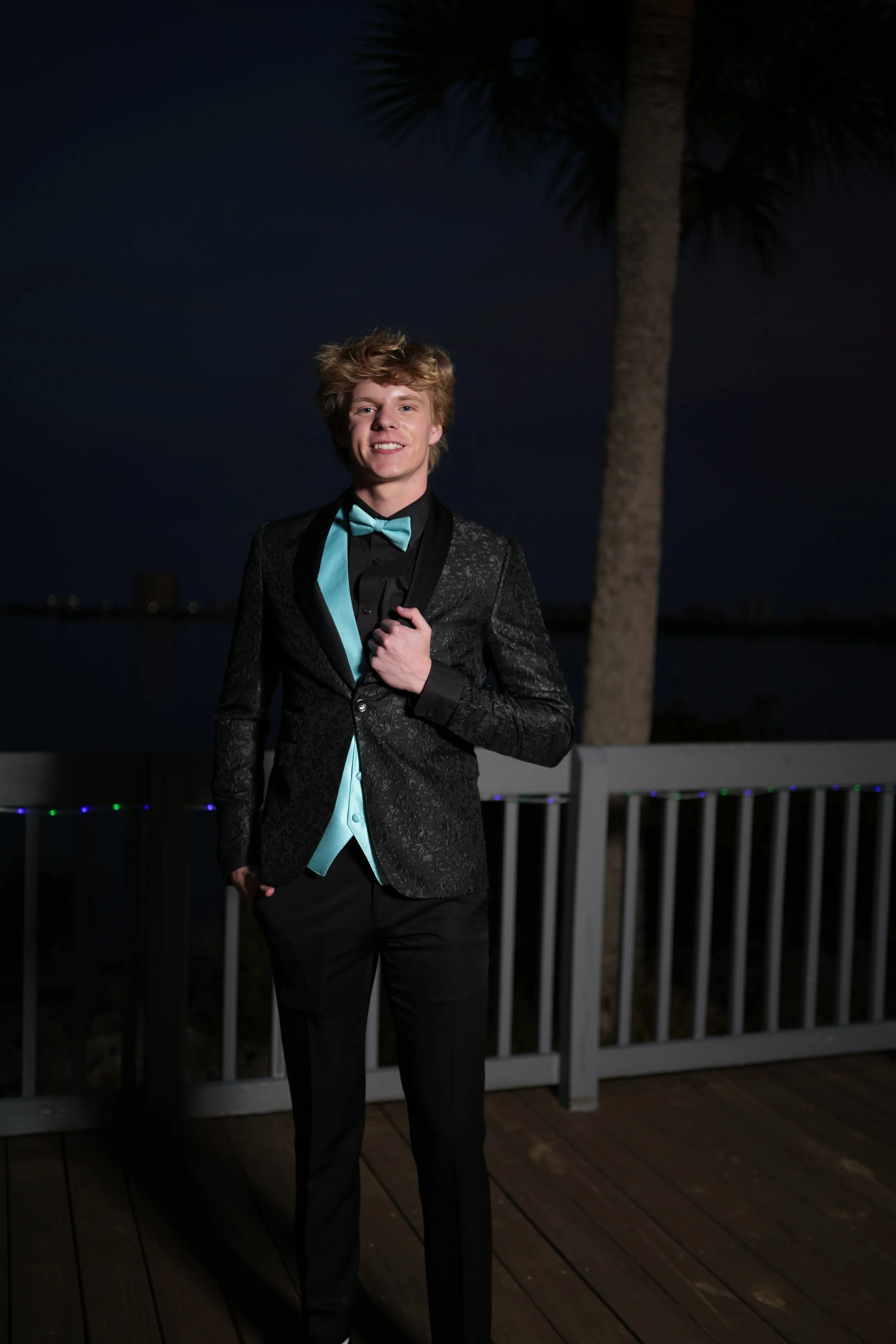 A young man in a black tuxedo with a turquoise bow tie and vest, standing outdoors at night on a wooden deck near a body of water with a palm tree in the background.