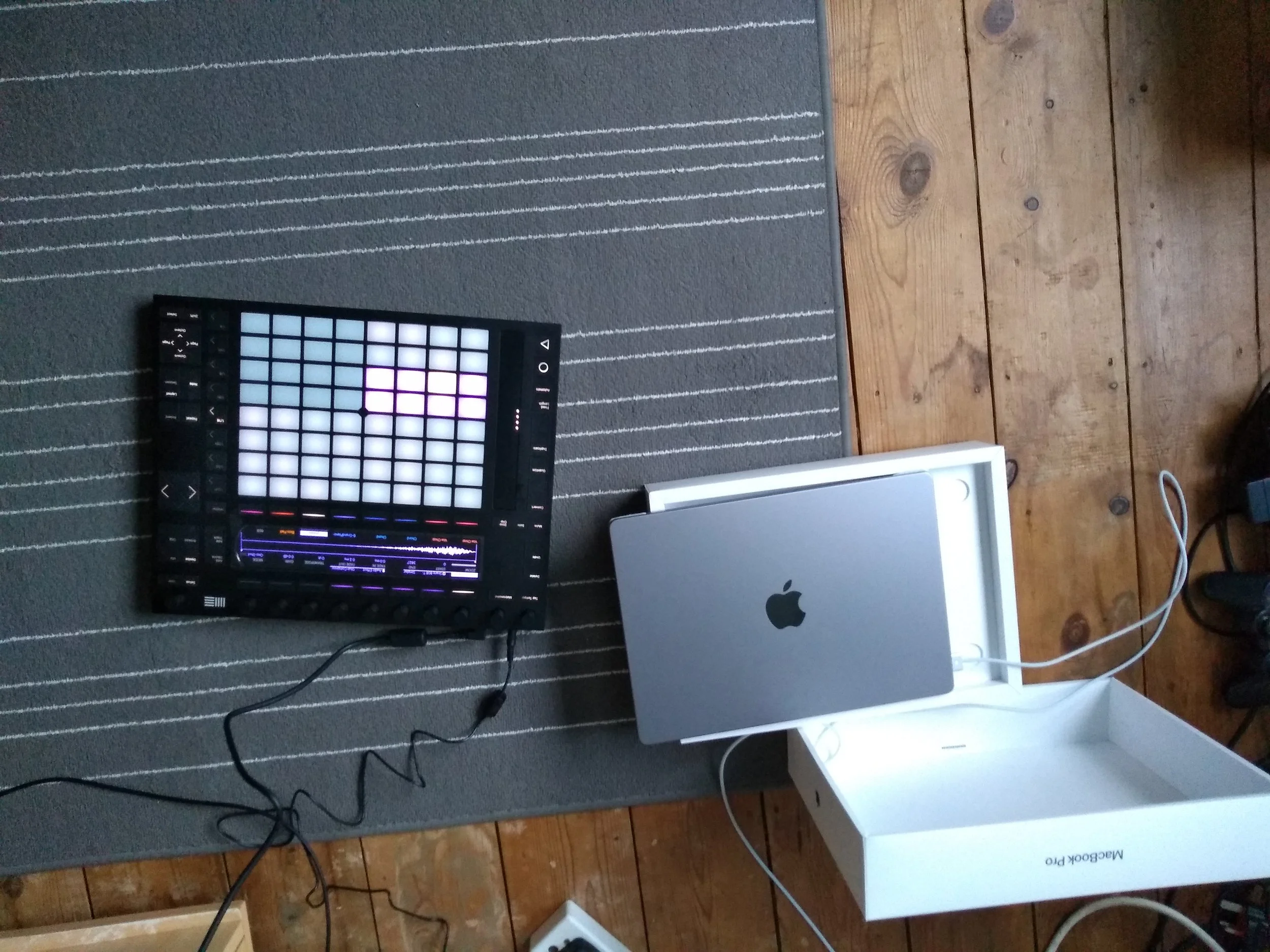 A music production setup with a Novation Launchpad connected to a MacBook Pro on a wooden floor and a striped rug.