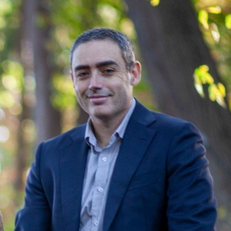Headshot of a man with short dark hair, blue eyes, wearing a checkered shirt, smiling softly against a brown and gray background.