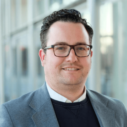 Professional man with dark curly hair, glasses, wearing a gray blazer, in an office environment.