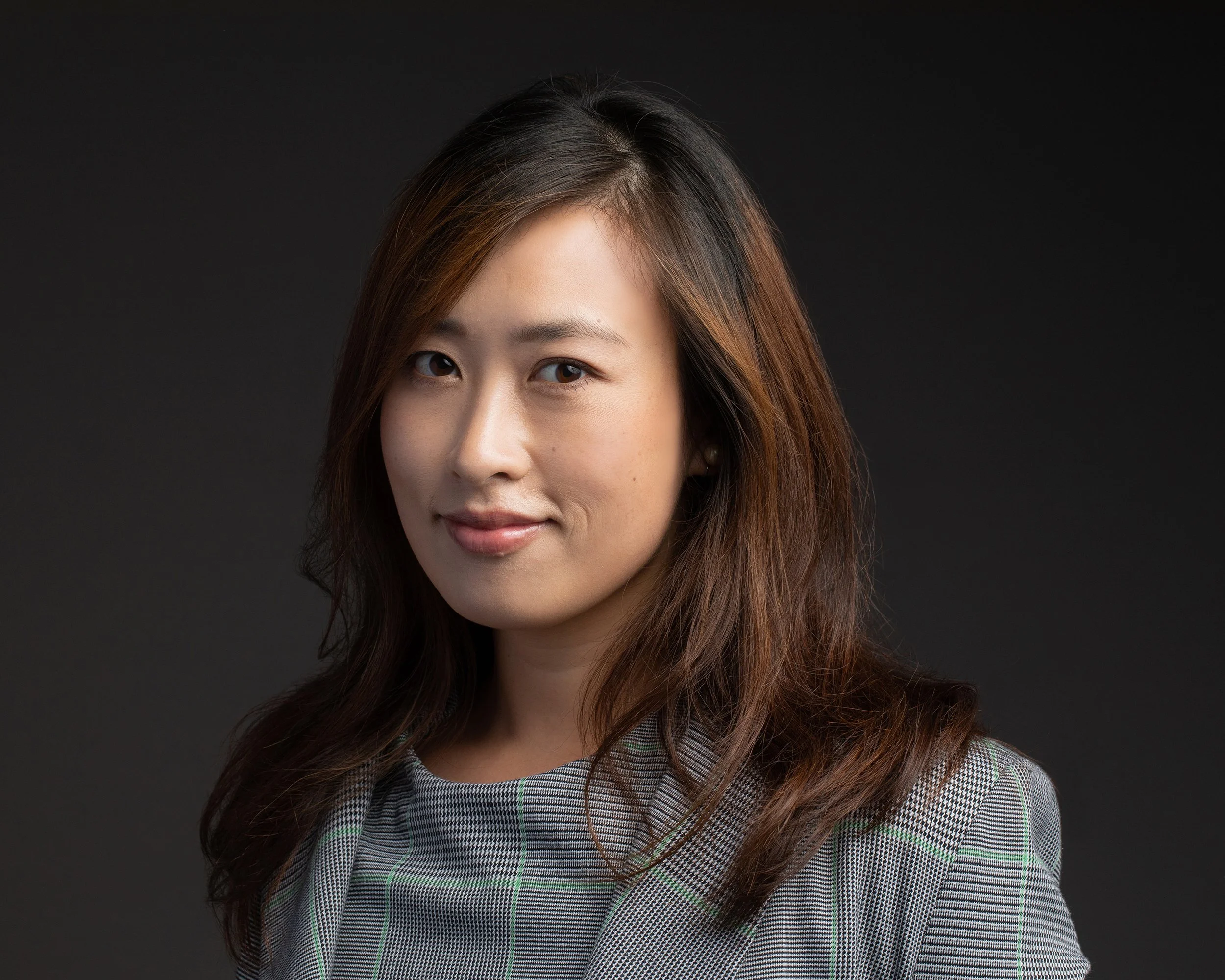 Portrait of a woman with shoulder-length brown hair, wearing a checkered blazer, against a black background.