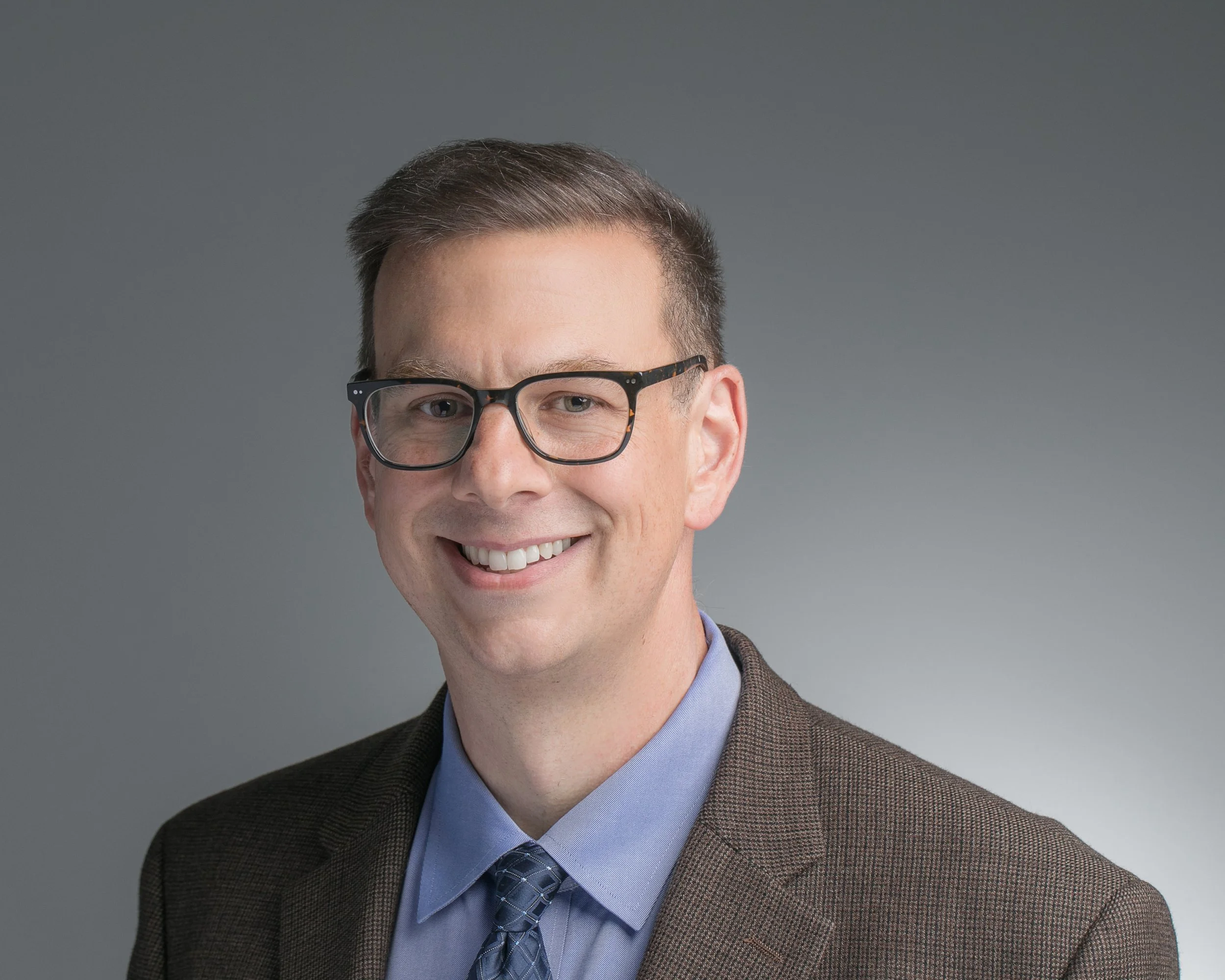 Headshot of a man wearing glasses, a brown blazer, a light blue shirt, and a patterned tie, smiling in front of a neutral background.