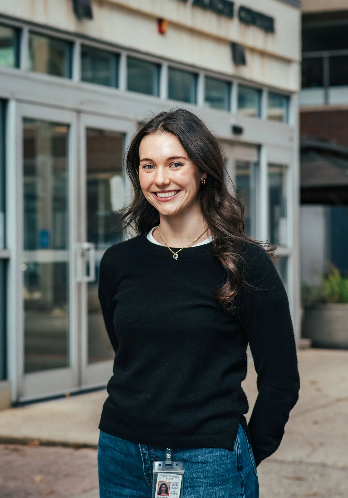 A woman with long dark hair and light skin standing outside a building with glass doors and windows, wearing a black sweater and blue jeans.