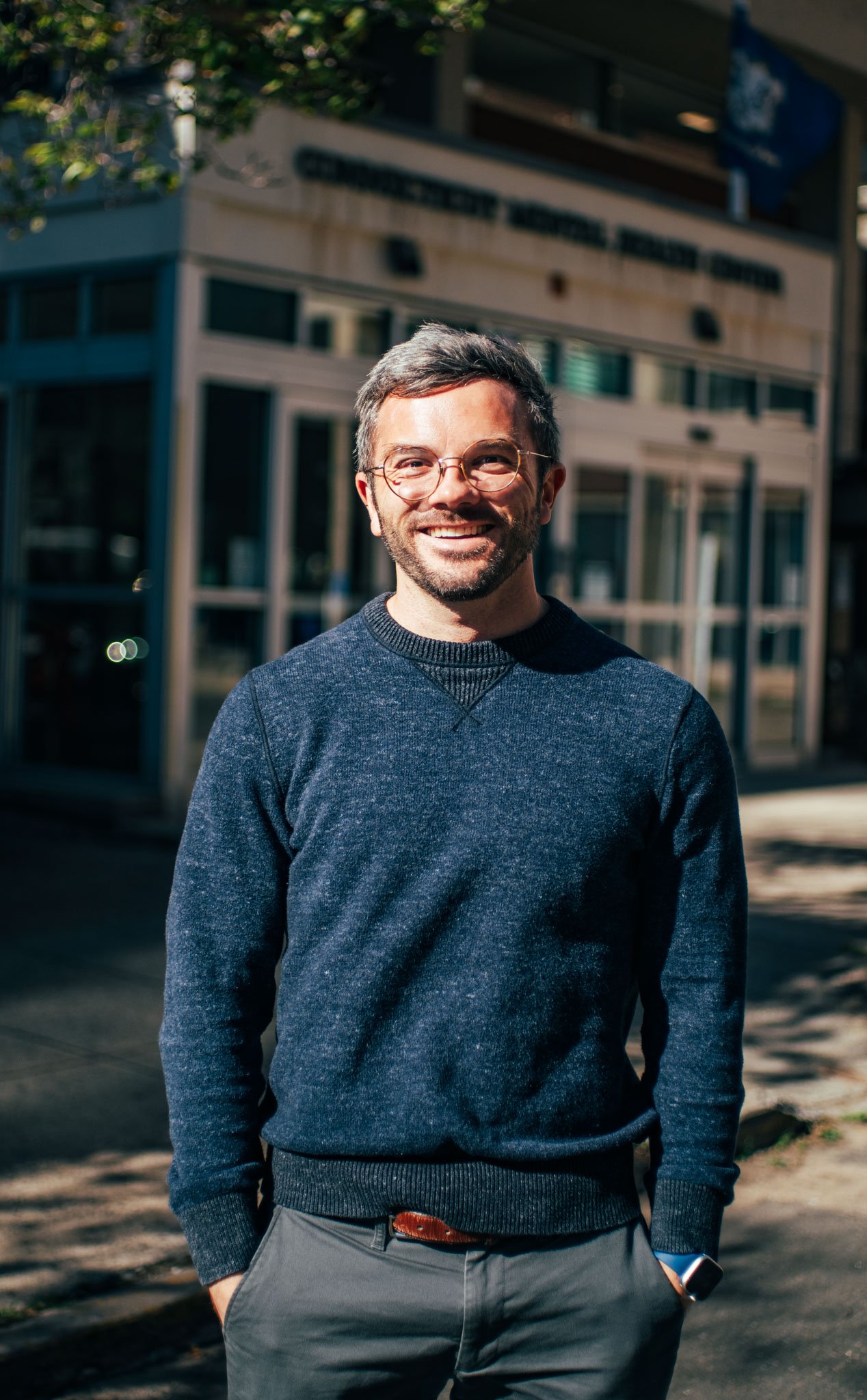 A man with glasses and a beard smiling outdoors on a sunny day, wearing a dark blue sweater and gray pants, standing in front of a building with glass windows.