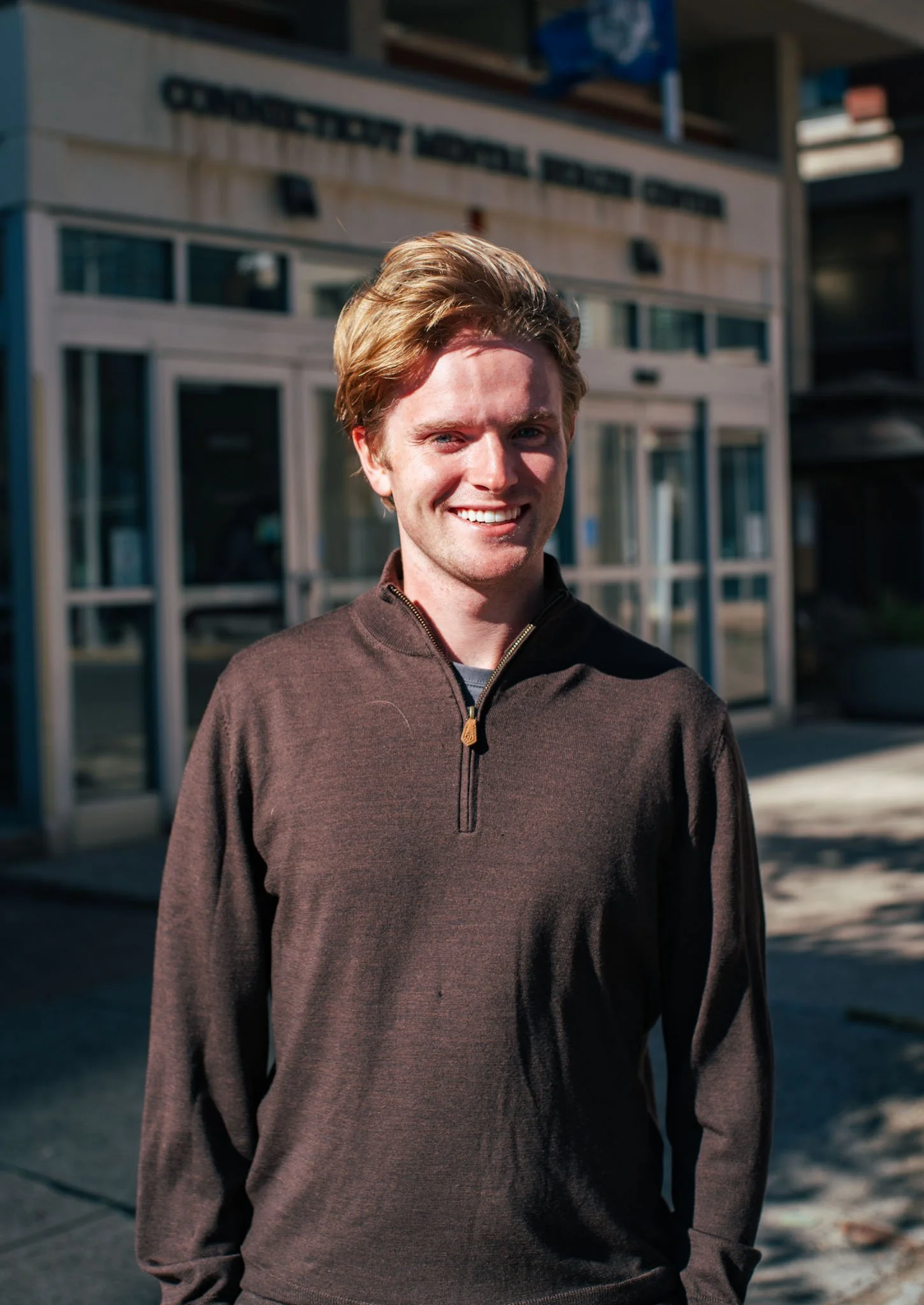 Young man with brown hair smiling outdoors in front of a building with glass doors.