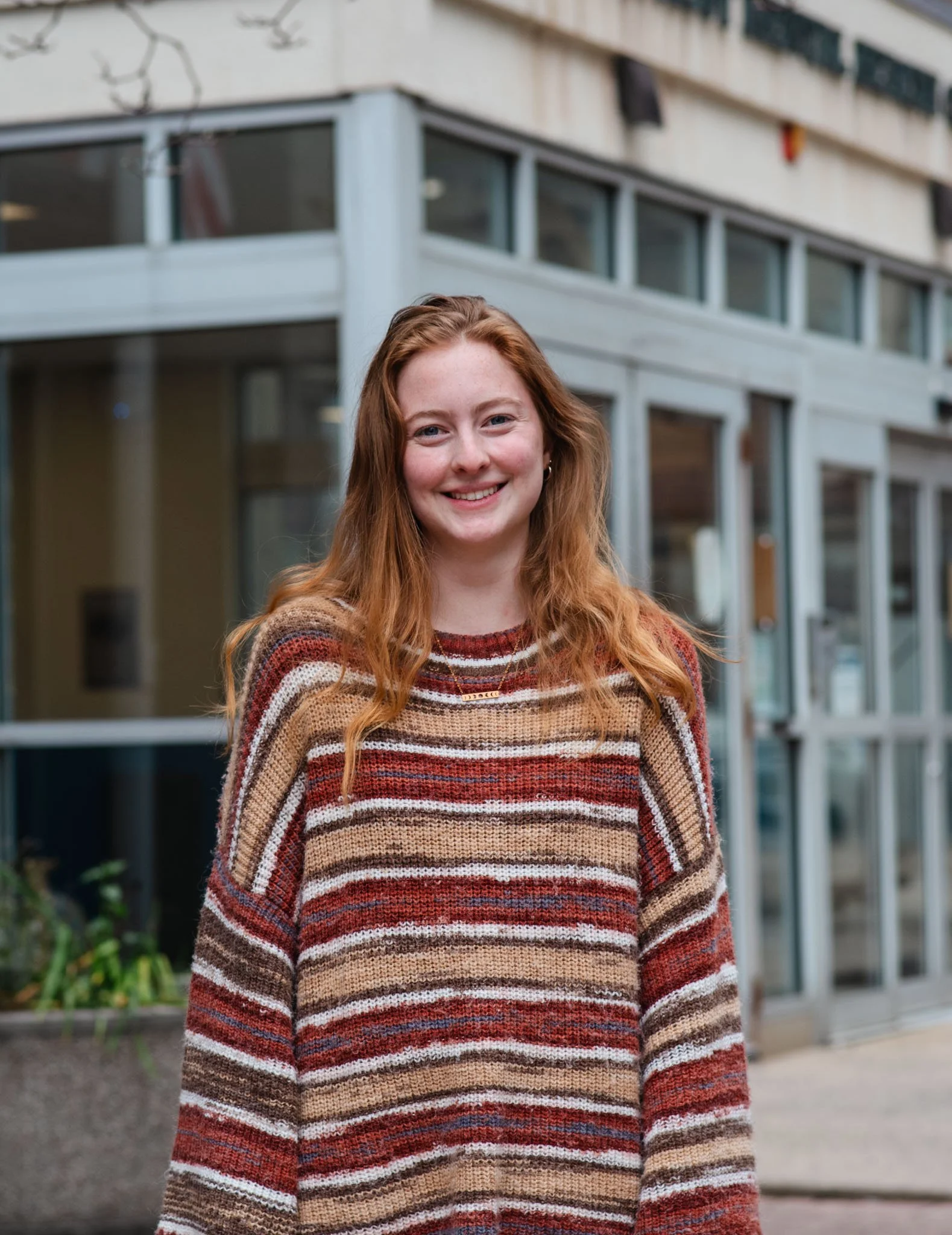 A young woman with long red hair and fair skin smiling outdoors in front of a modern glass building.