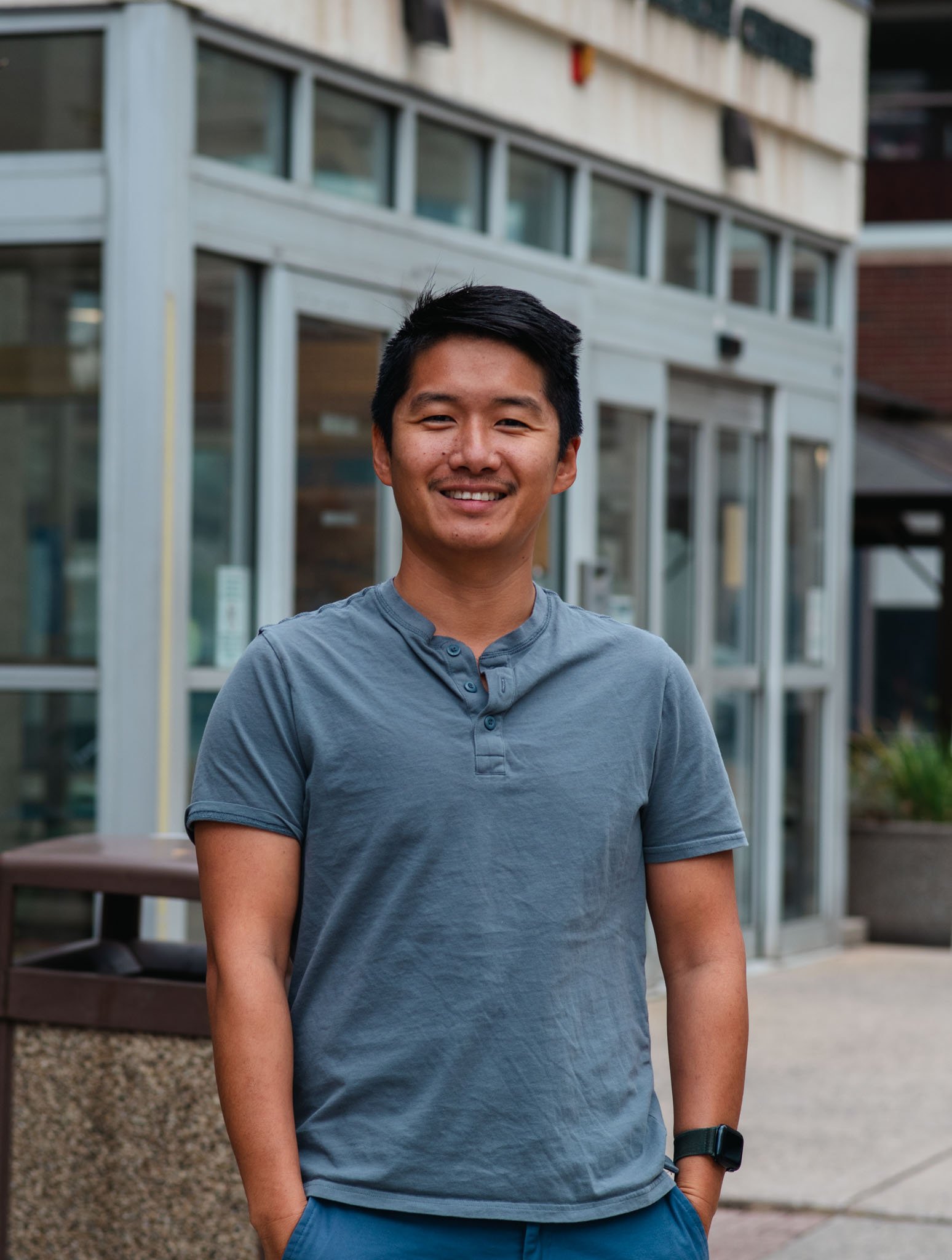 Portrait of a young Asian man in a navy suit and light blue shirt, smiling outdoors in front of a blurred modern brick building.