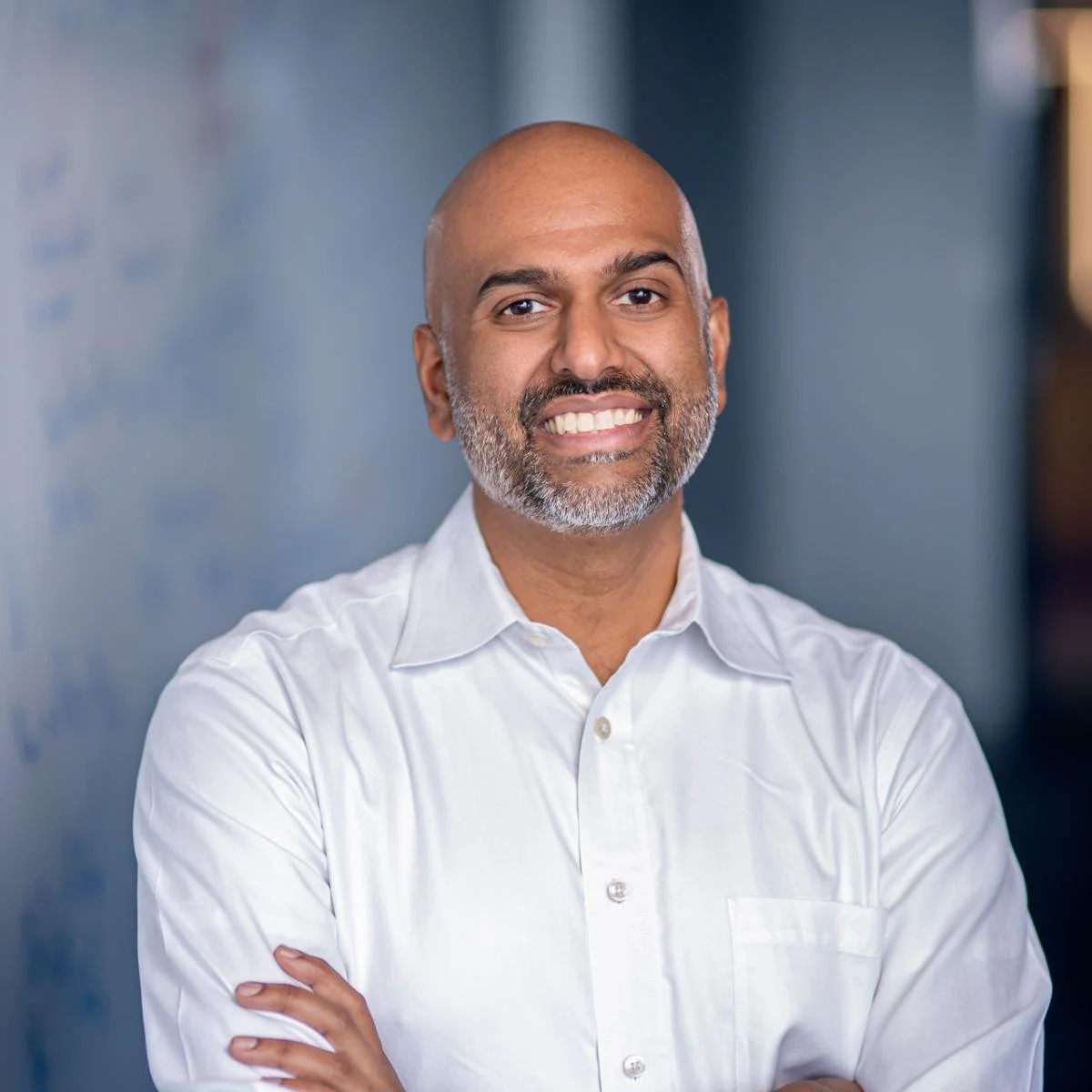 A smiling man with a beard and bald head, wearing a white button-down shirt, standing with arms crossed in an office setting.