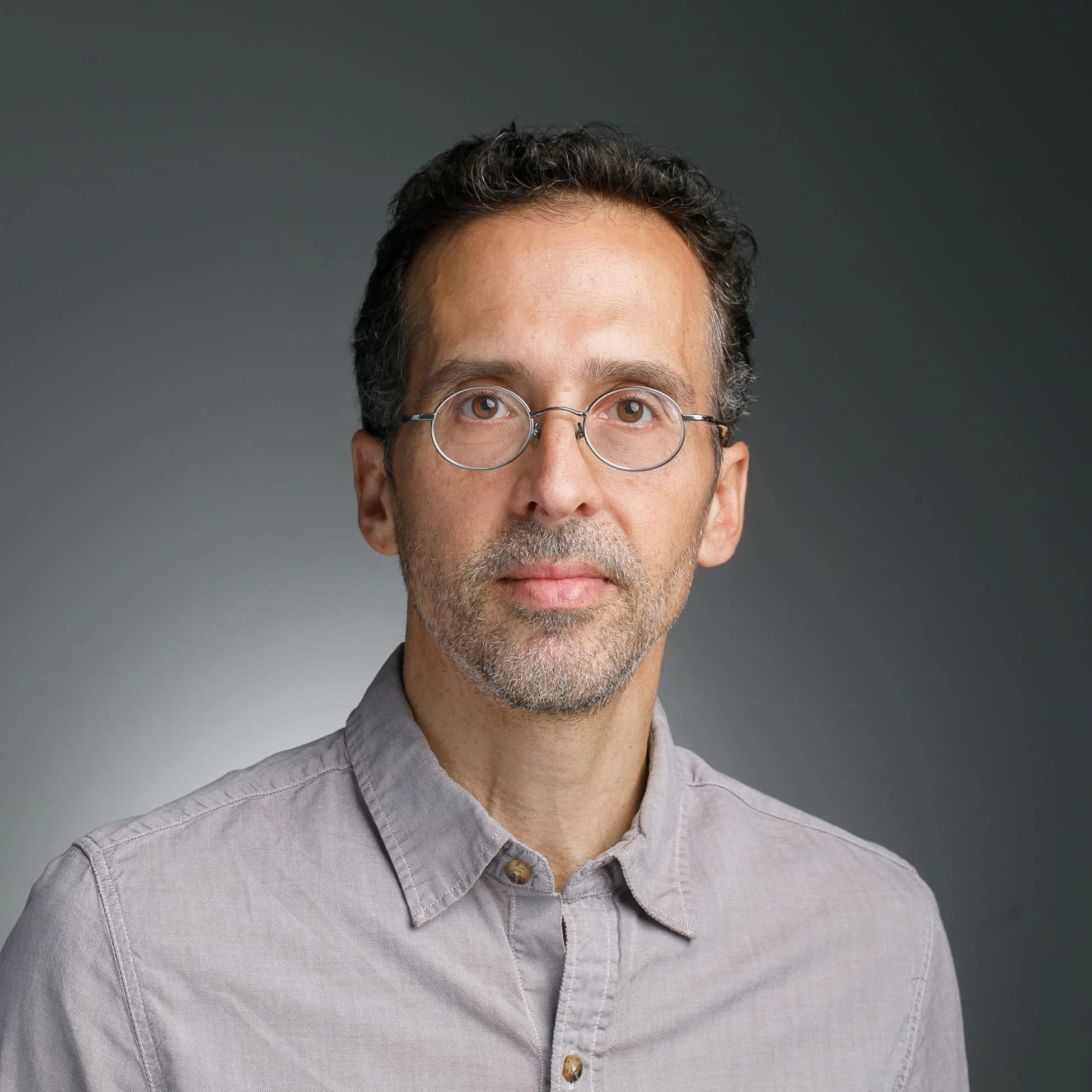 Headshot of a middle-aged man with dark hair and glasses, wearing a light gray button-up shirt, against a dark gray background.