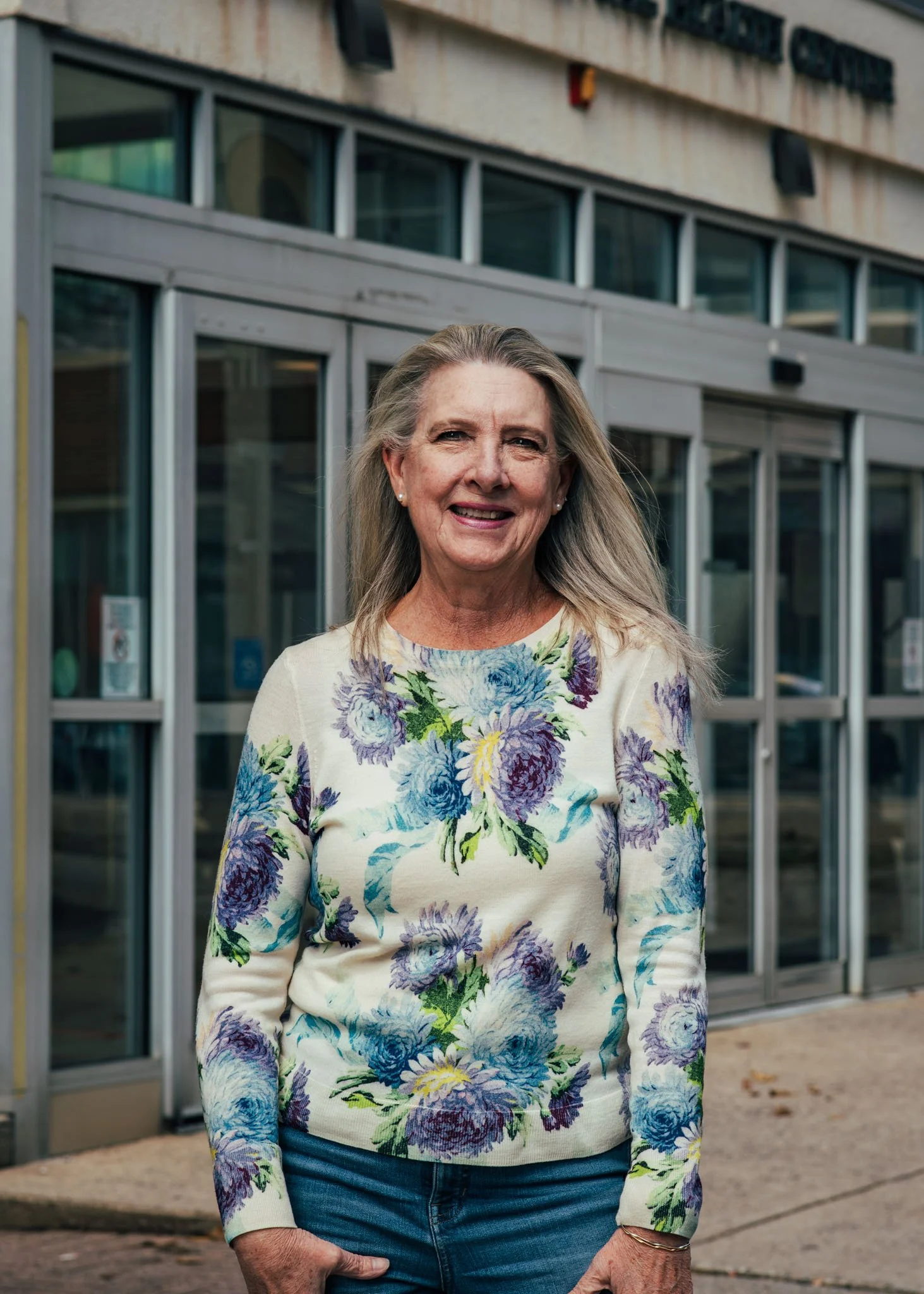 An older woman with long gray hair standing outside in front of a building with glass doors and windows, wearing a floral sweater with shades of blue, purple, and green.