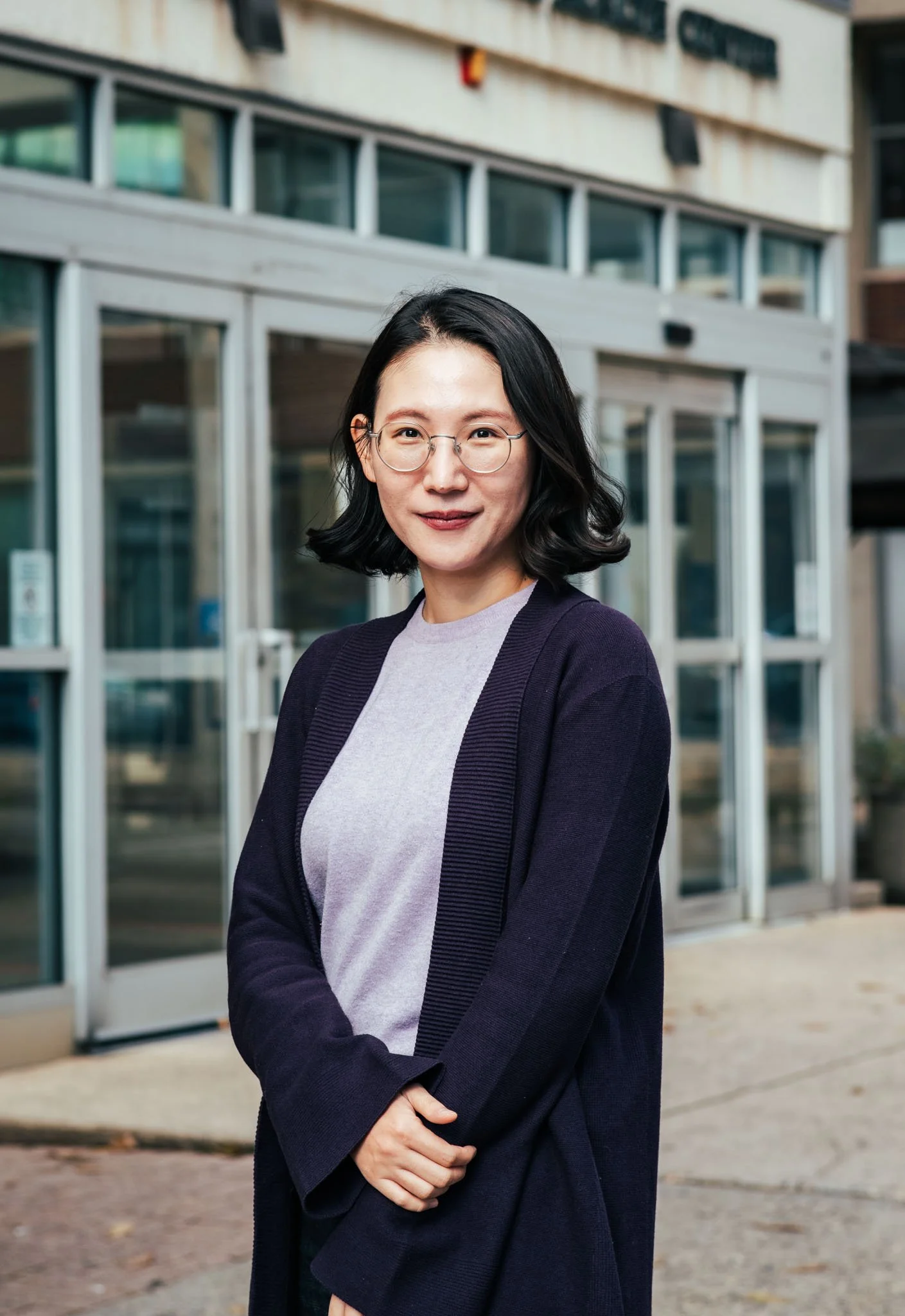 A woman with glasses and shoulder-length black hair smiling outdoors in front of a building with glass doors and windows.