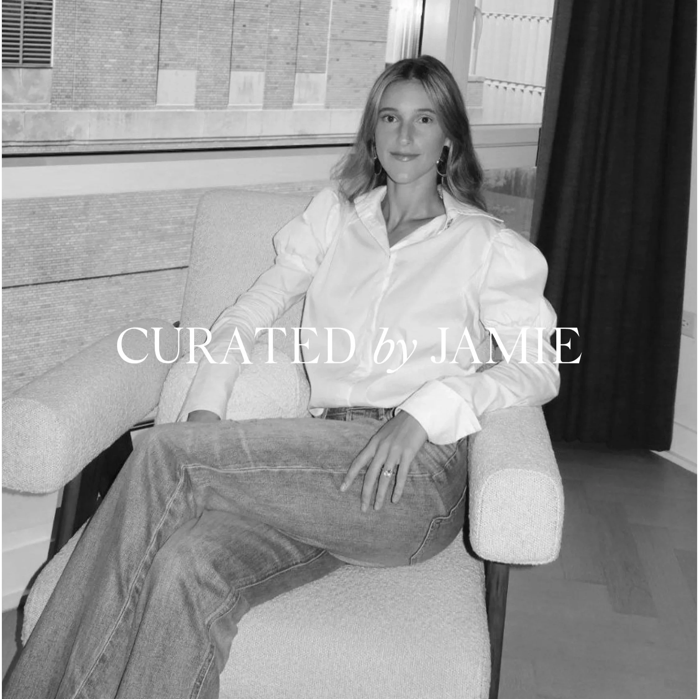 A woman with long hair sitting on a sofa in front of a window. She is wearing a white shirt and jeans. Behind the window, there is a brick building.