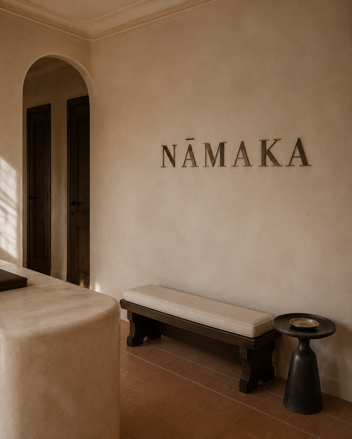 Interior with a beige wall decorated with metallic letters spelling 'NāMĀKA', a black wooden bench with a beige cushion, and a black side table with a small dish, bamboo tile flooring, and wooden doors.