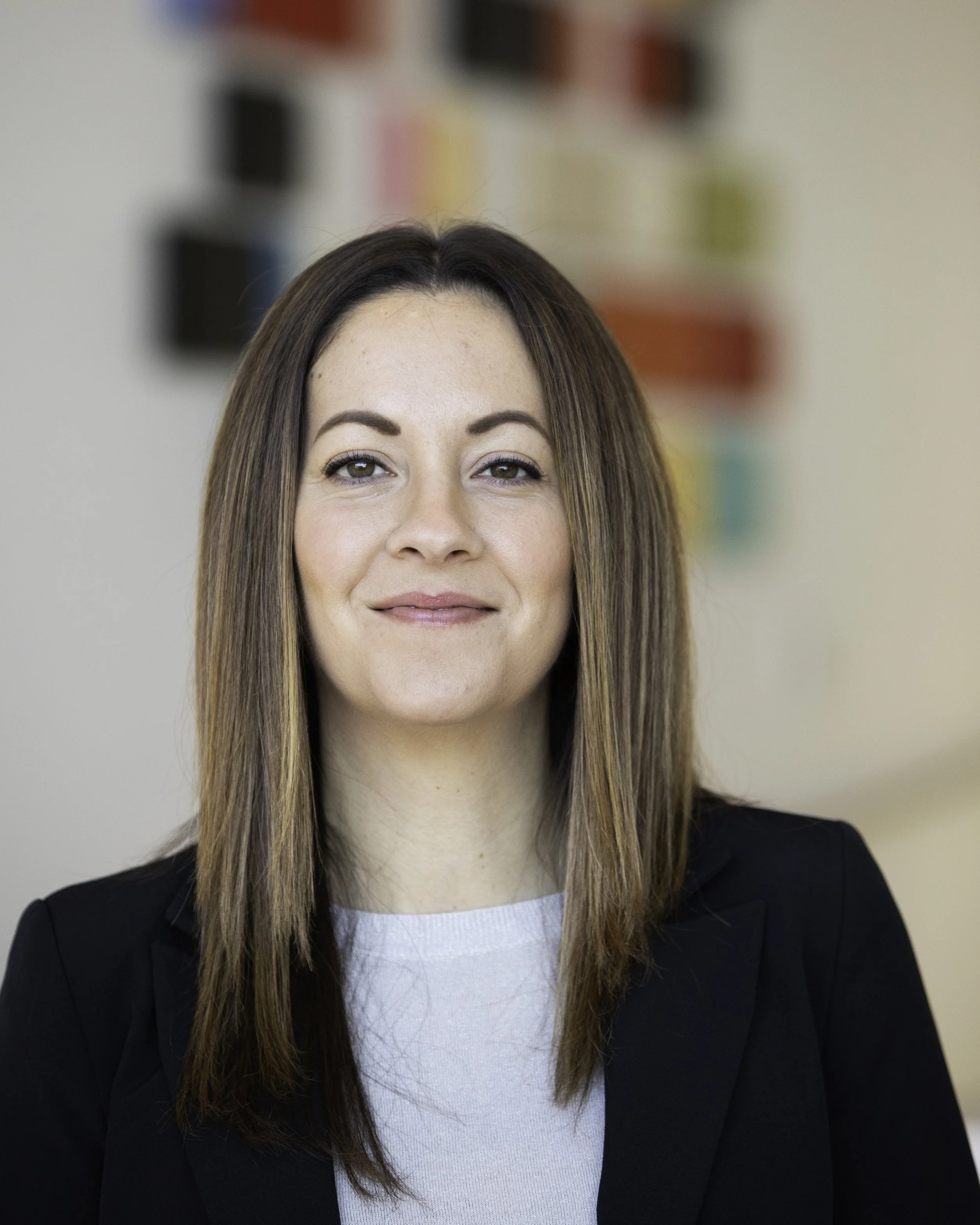 A portrait of a smiling woman with long brown hair, wearing a blue patterned shirt and a dark blazer, standing outdoors with a blurred green background.
