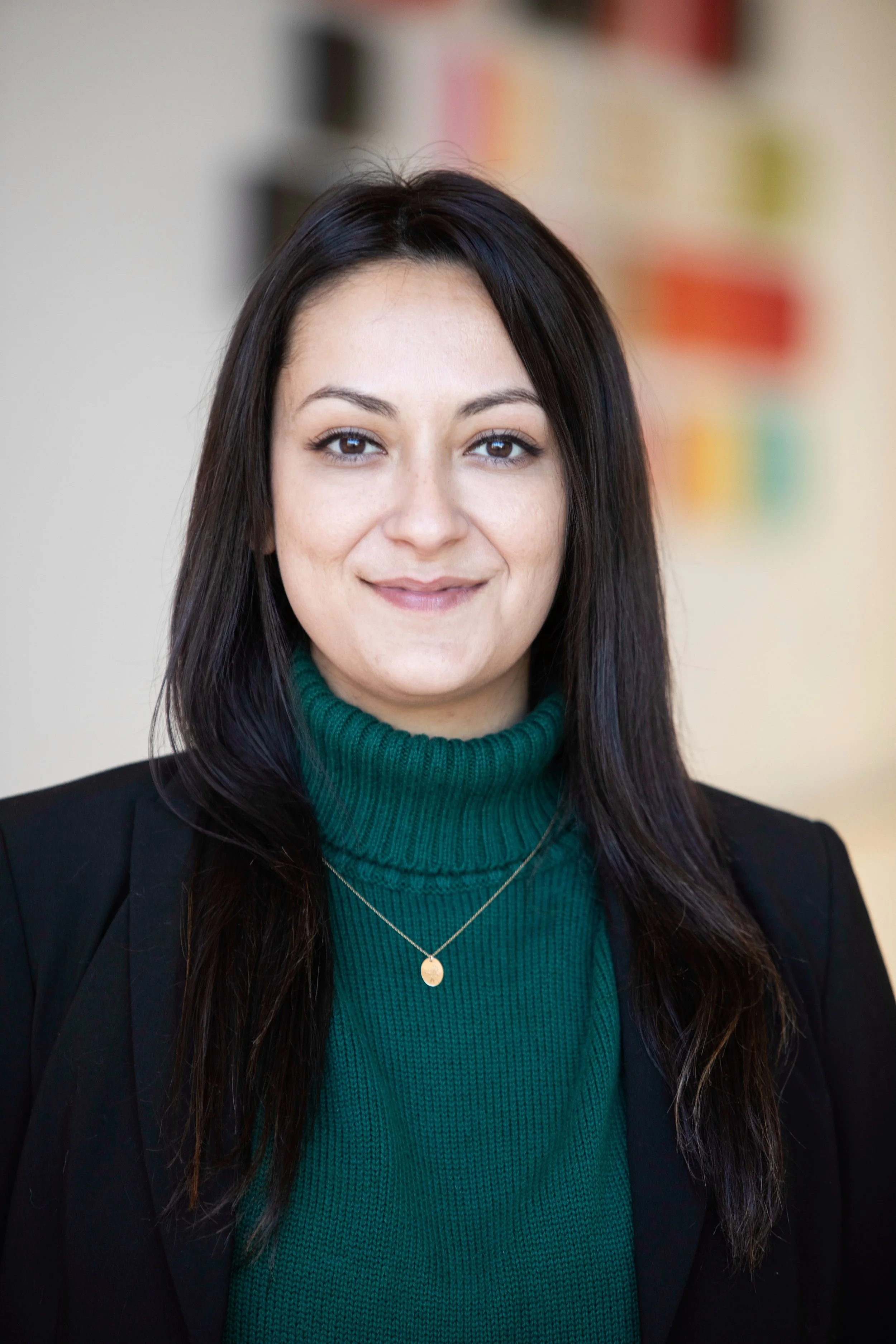 A woman with long, dark, curly hair and light skin, smiling at the camera against a plain gray background.