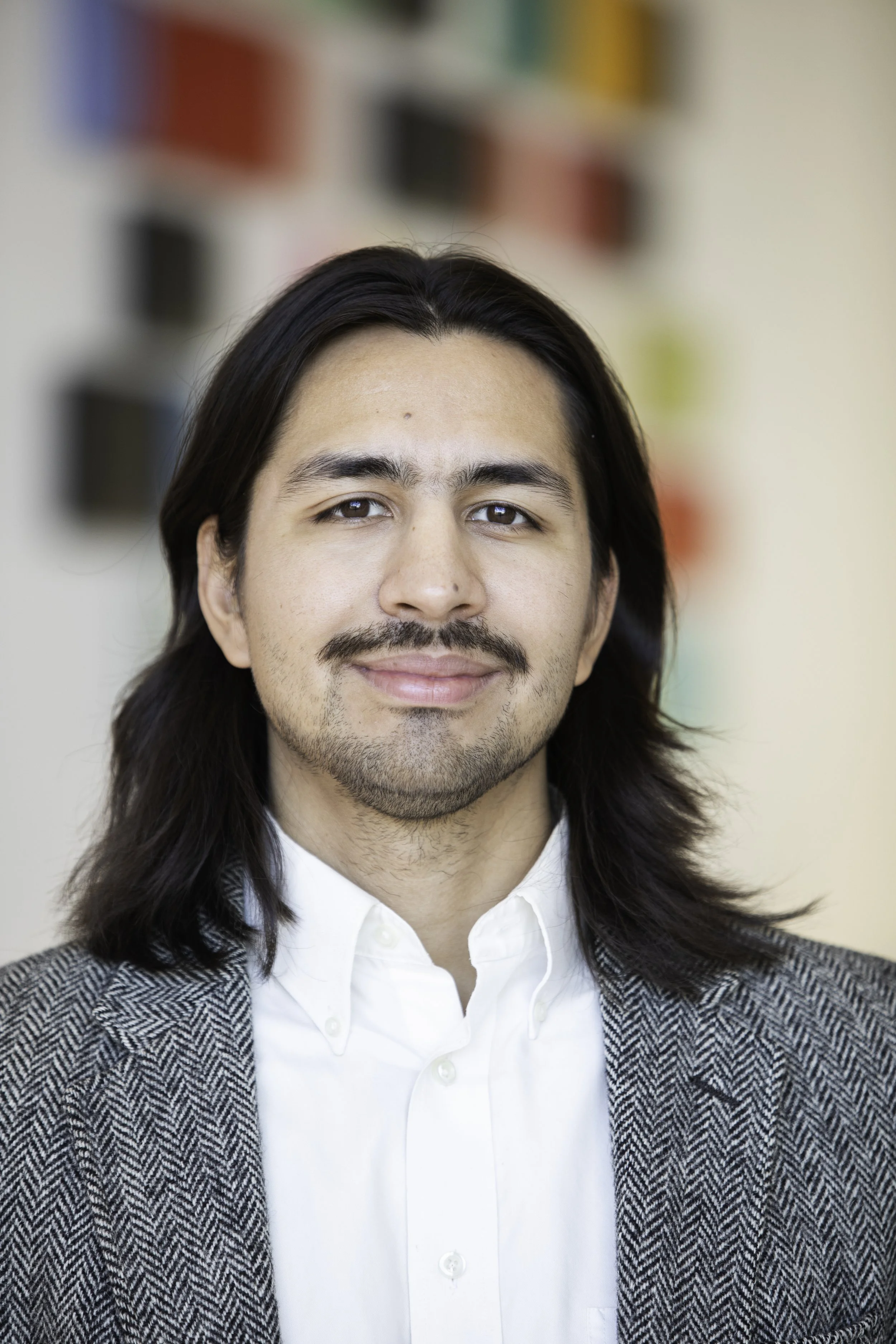 A young man with long dark hair, smiling, wearing a black turtleneck and a gray blazer, indoors in a well-lit environment.