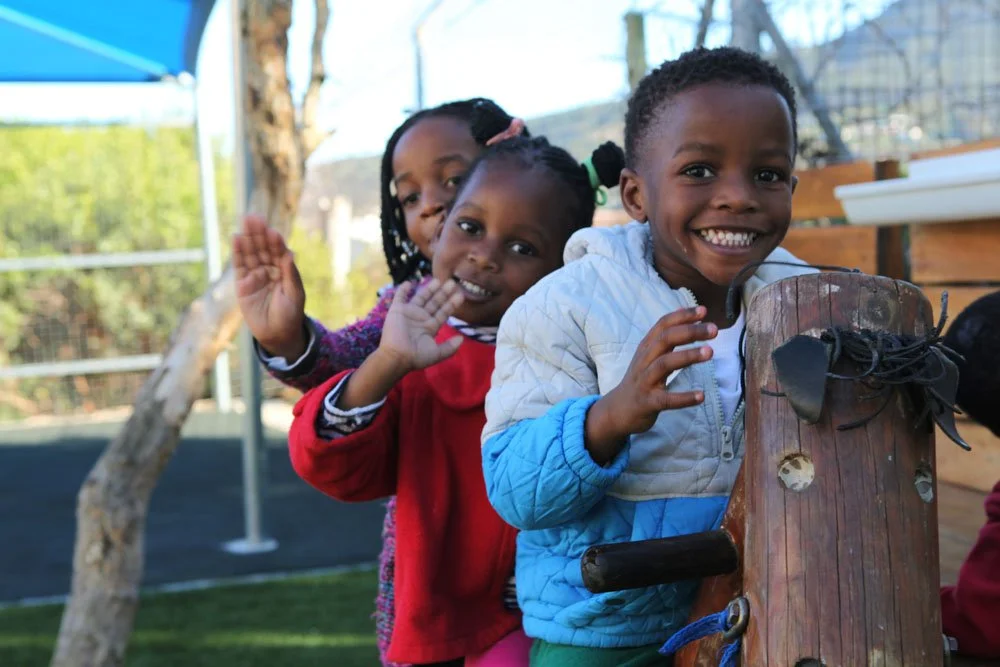 Kinder smilen und winken von einem Holzpferd sitzend in die Kamera. Sie sind auf dem Spielplatz von Little Lambs bei Kapstadt.
