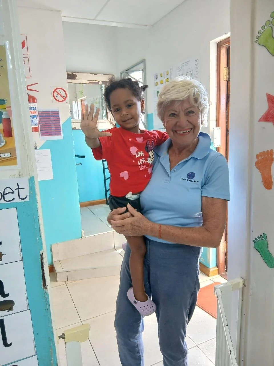 Elke Zwicker, Trustee von The Kinder Hilfe Kapstadt trägt ein winkendes Kind auf dem Arm.
