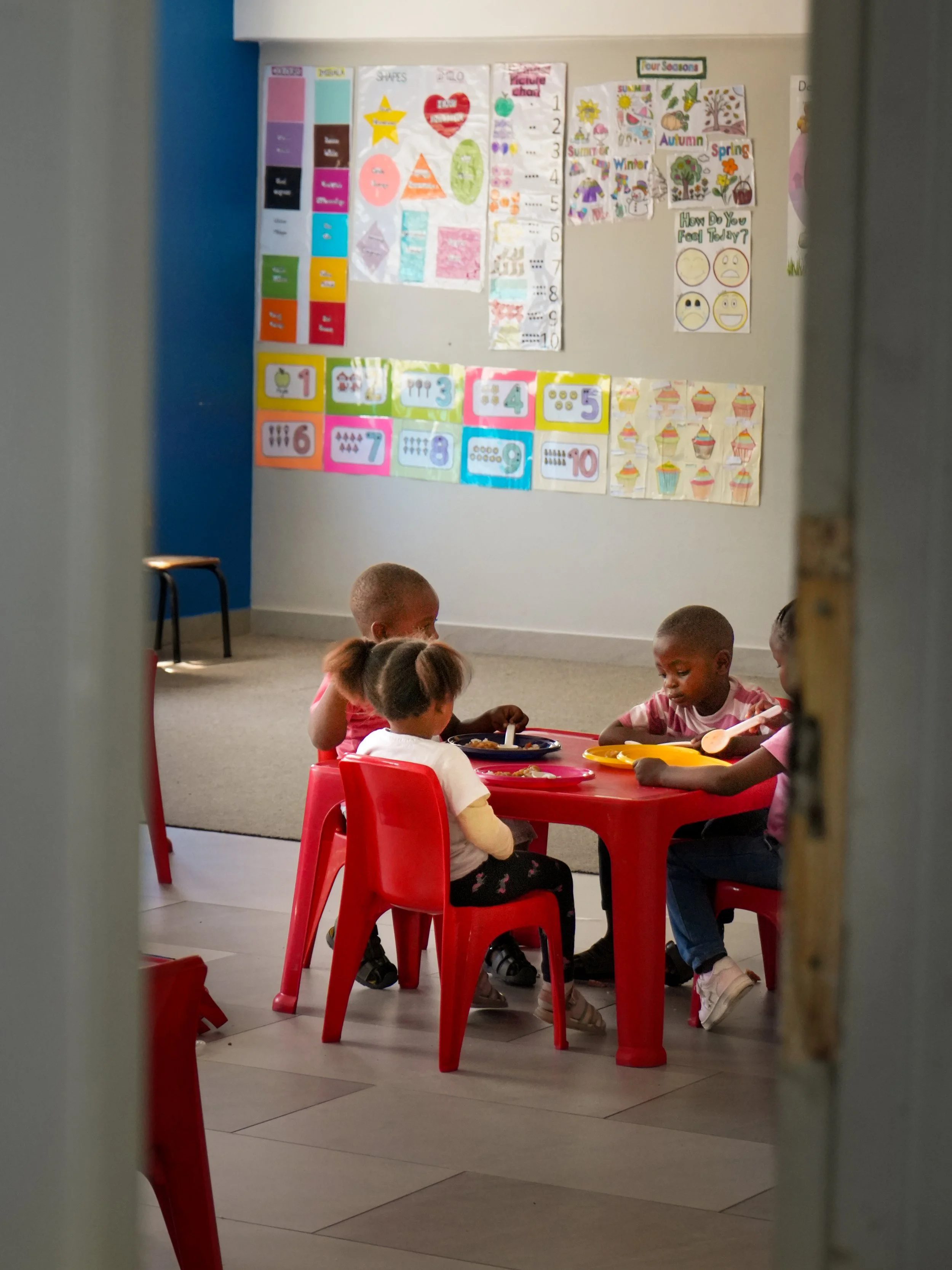 Kinder essen gemeinsam zu Mittag auf ihren Plastikstühlen an einem Plastiktisch.