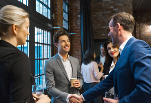 A group of professionally dressed people at a social gathering, shaking hands and engaging in conversation indoors near large windows.