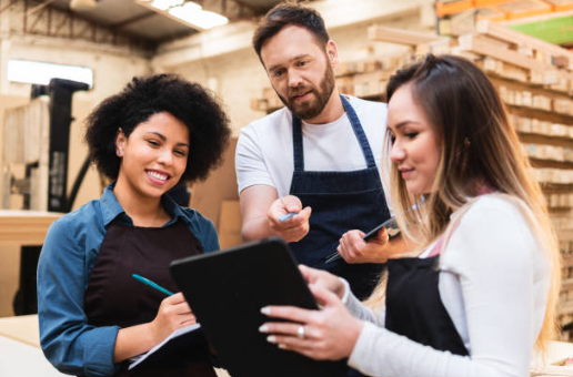 Three people in a workshop or craft store, one man and two women, looking at a tablet, wearing aprons, surrounded by wooden shelves and wood pieces.