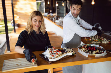 A woman and a man preparing desserts in a modern kitchen with large windows and natural lighting.