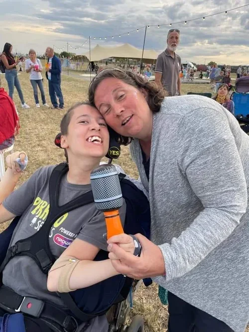 A young girl in a wheelchair holding an inflatable microphone, smiling, with an adult woman leaning over her at J-Fest with the festival in the background.