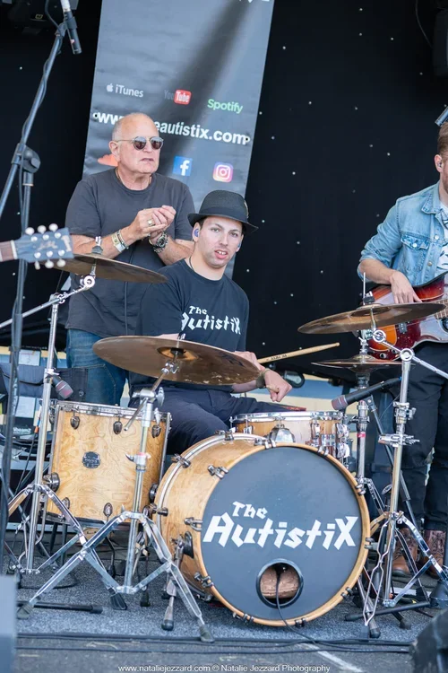 Three musicians performing on stage, with a drum set labeled 'The AutistiX' in front of them. One man is playing drums, another man is playing guitar, and a third man is standing behind the drummer.