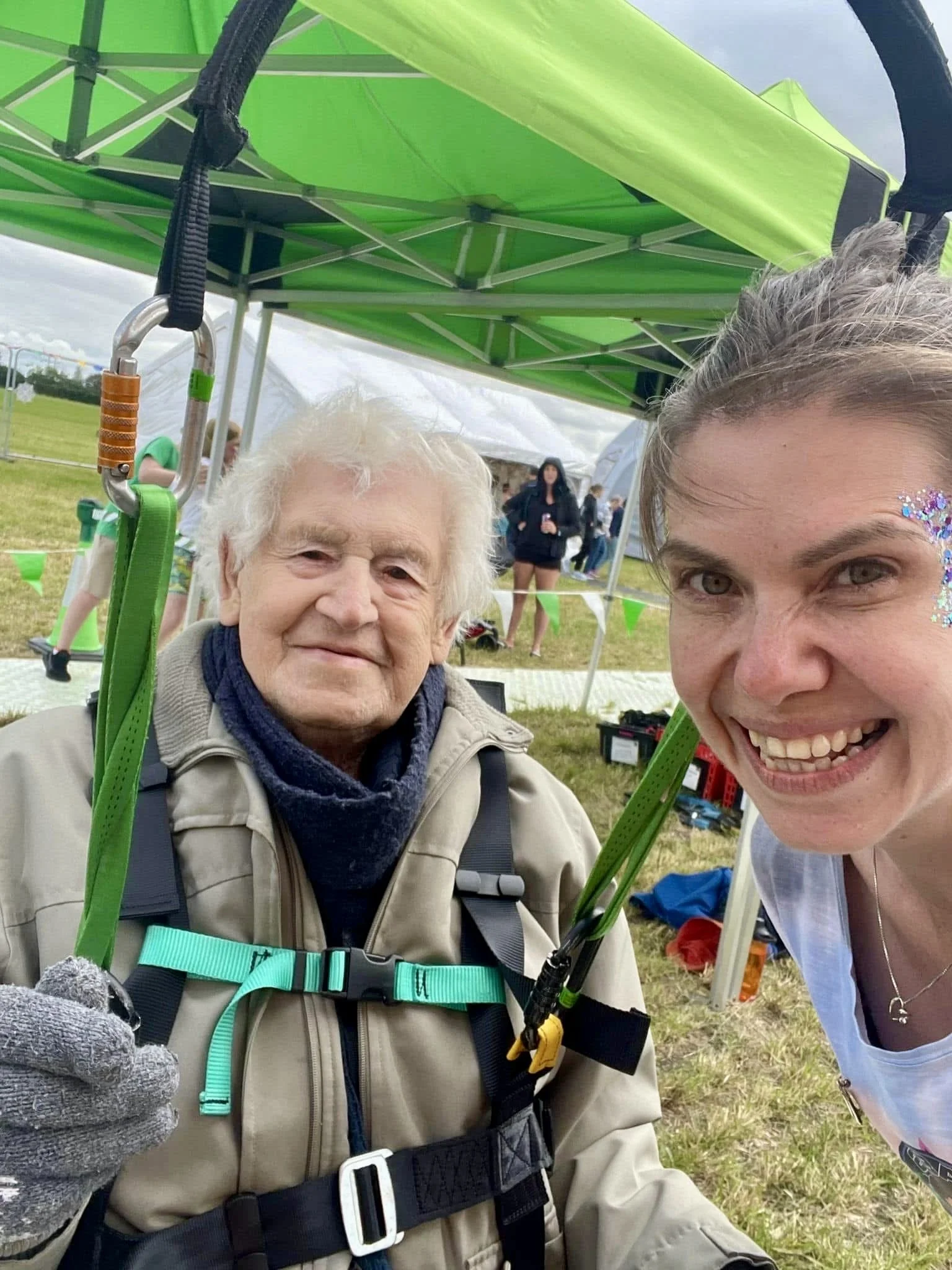 A woman and an elderly man smiling for a photo at an outdoor event under a green canopy tent. The man is wearing a beige jacket, blue scarf, and gloves, with a harness and climbing gear.