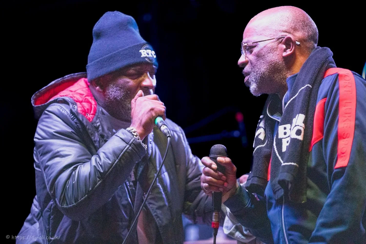Two men on stage, one wearing a black beanie and gray jacket, and the other with glasses and a jacket with a towel around his neck, are holding microphones close to each other, seemingly in a conversation or performance.