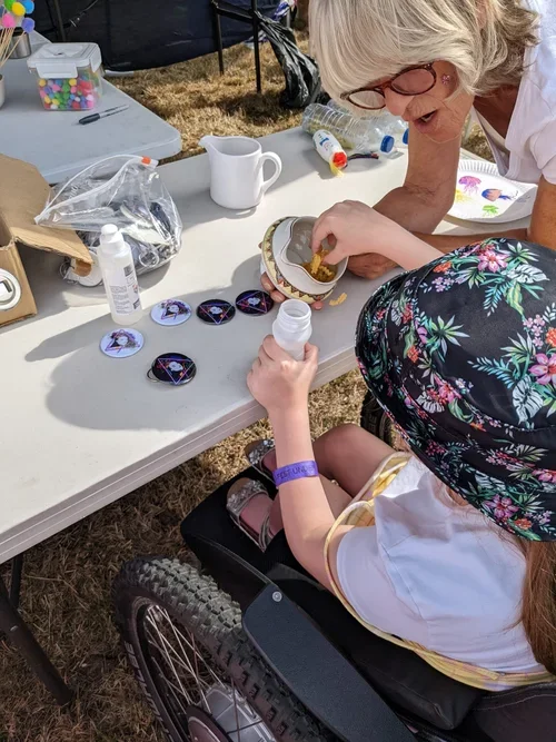 A woman assisting a young girl in a wheelchair with crafting at a table outdoors. The table has various decorating supplies.