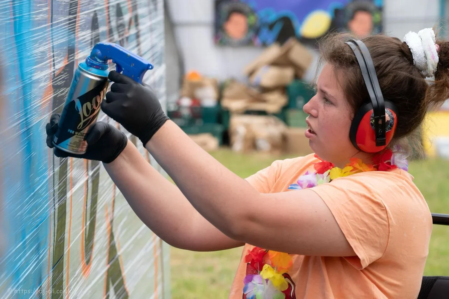 A young girl in a wheelchair, wearing ear defenders, a colourful orange shirt is spray painting on a wall with a can of spray paint.
