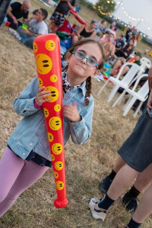 A young girl with glasses and braided hair holding a large red inflatable baseball bat decorated with yellow smiley face emojis at festival with people sitting at tables and string lights overhead.