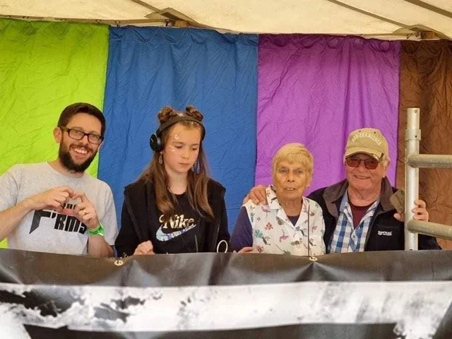 Four people standing behind a DJ booth with a colorful backdrop of green, blue, purplecurtains. The group includes a young man with glasses making a heart gesture, a young woman wearing headphones - she is the DJ - an older woman, and an older man.