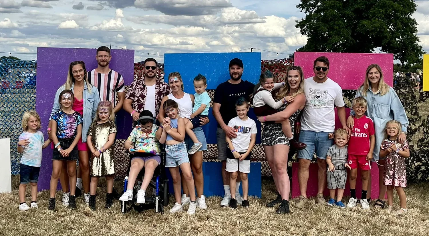 Group of adults and children both standing and in wheelchairs, posing outdoors in front of colourful J-FEST sign, with some children and adults smiling and interacting, on a grassy field with a cloudy sky overhead.