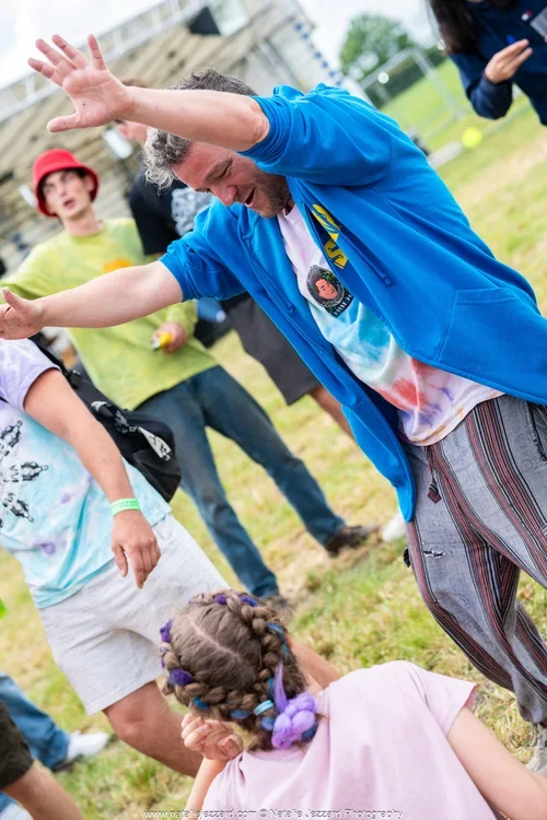A man with curly gray hair wearing a blue jacket and patterned pants is bending forward with arms outstretched, engaging with a young girl with braided hair in a pink shirt. Several people are in the background on a grassy field, some watching and others standing, with a building and trees in the distance.