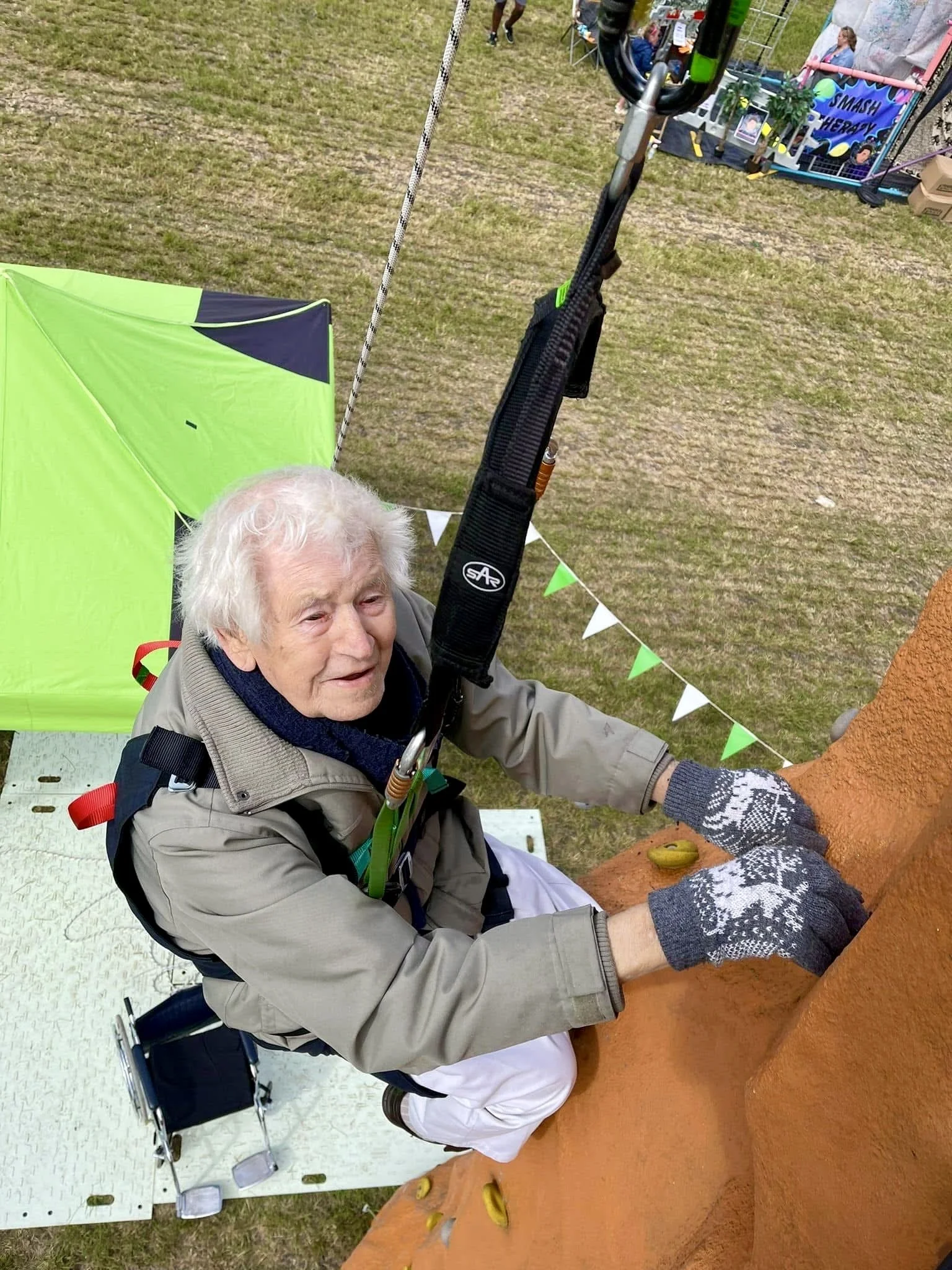 An elderly man with white hair rock climbs in a harness, and is smiling while climbing an orange rock wall at an outdoor event, with a green tent and some people in the background.