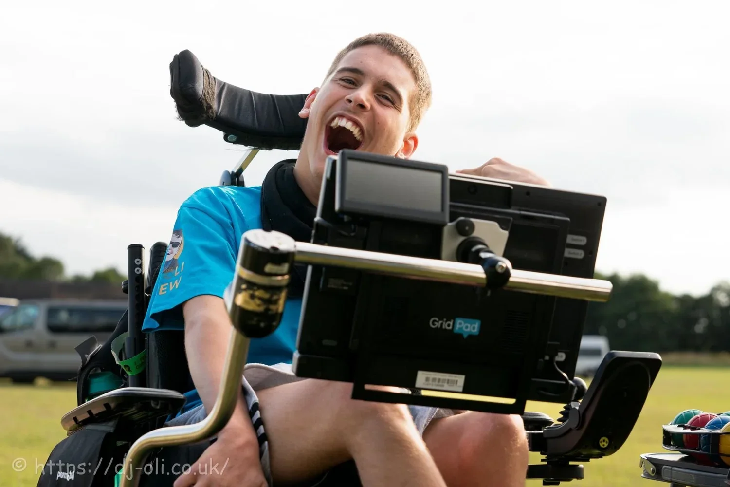 Young man in a wheelchair outdoors, smiling and laughing, with computer screen he controls only with his eyes to communicate. He is at J-Fest.