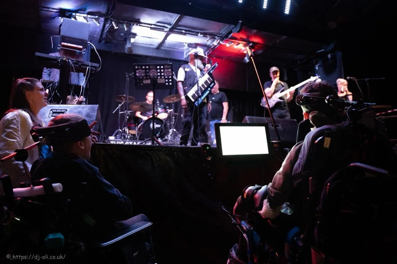 A band performing live on stage with a drummer, keyboardist, bassist, and guitarist. Audience members include people in wheelchairs watching the performance.