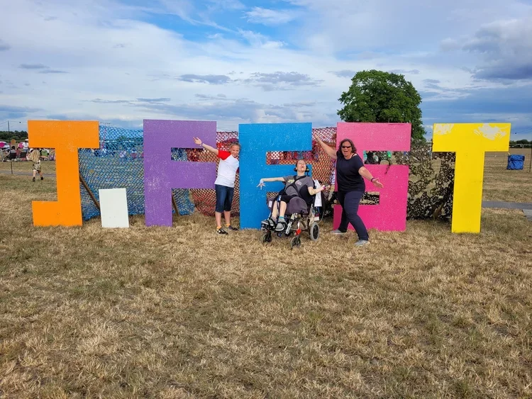 People standing in front of large, colourful letters spelling 'J-FEST' outdoors, with grassy field and cloudy sky in the background.