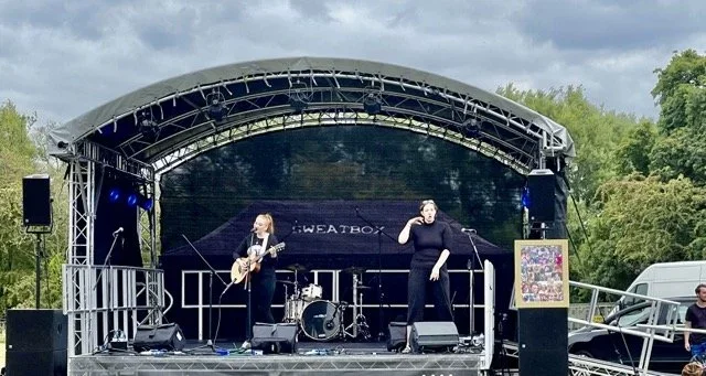 A live outdoor music performance on a stage with one female singer and one BSL interpreter. There is a guitarist, and a drum set, under a curved canopy.
