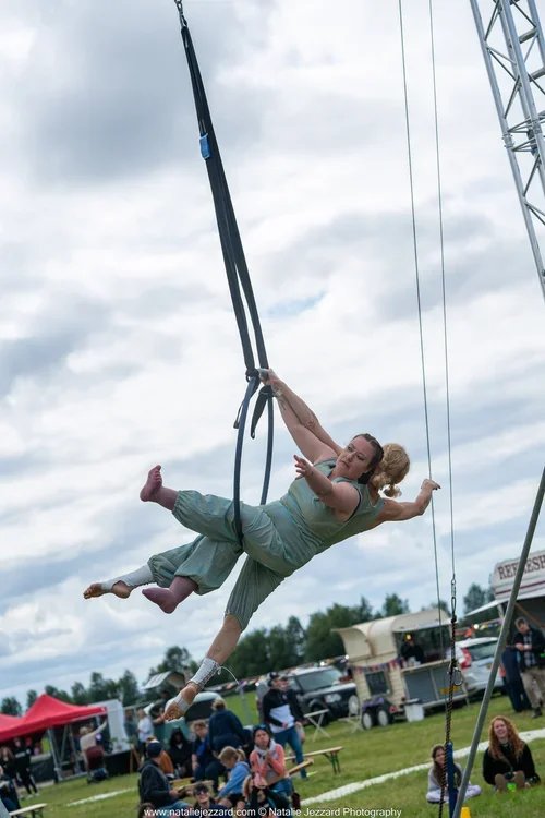 Two women are performing an aerial silk act outdoors at a fair or festival, with one woman supporting the other as she hangs from long fabric strips, and a cloudy sky overhead.