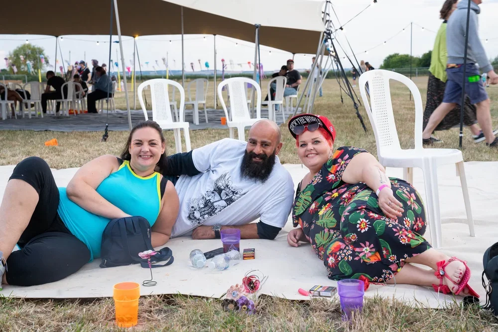 Three people lying on a white mat outdoors at a festival or outdoor event, smiling at the camera. A large tent is in the background, with white chairs and people sitting underneath. There are water bottles, cups, and small items on the mat, and some people walking or sitting in the background.
