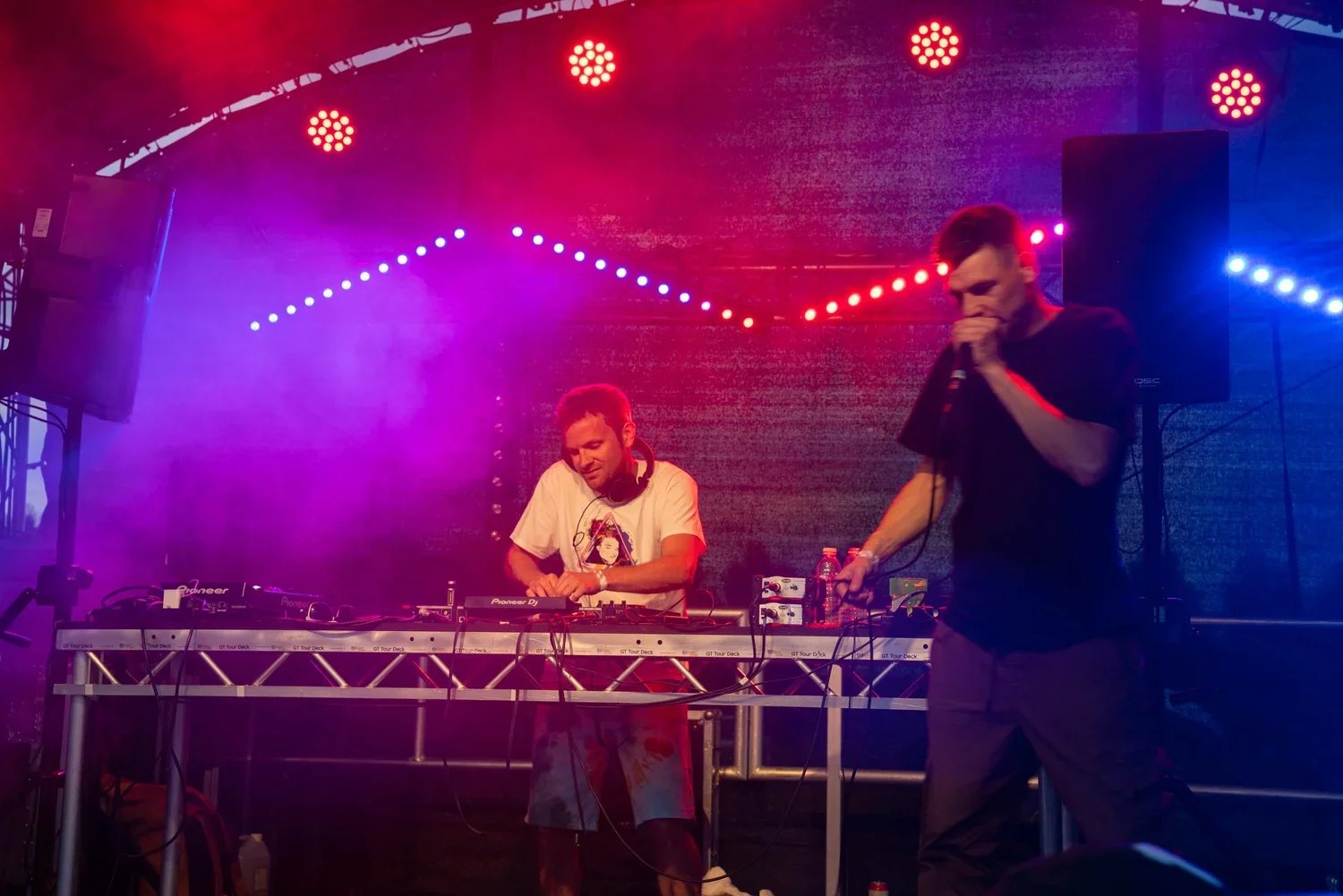 A DJ and an MC performing on stage with colourful red and purple lighting, the DJ wearing a white t-shirt with a J-FEST logo and the MC in a black t-shirt, with DJ equipment and water bottles in front of them.