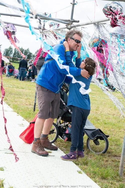 A man and a young girl are sharing a joyful moment under a colorful kite display at an outdoor festival. The man is wearing sunglasses, a blue jacket, and shorts, while the girl is dressed in a blue shirt and jeans. They are smiling and embracing, with a stroller nearby, and other festival attendees in the background.