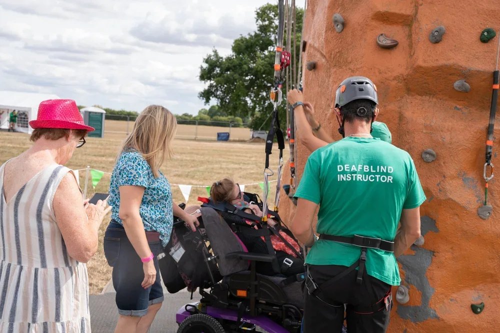 A deafblind instructor in a green shirt and helmet assisting a young girl in a wheelchair climbing a rock wall outdoors, with two women observing nearby.