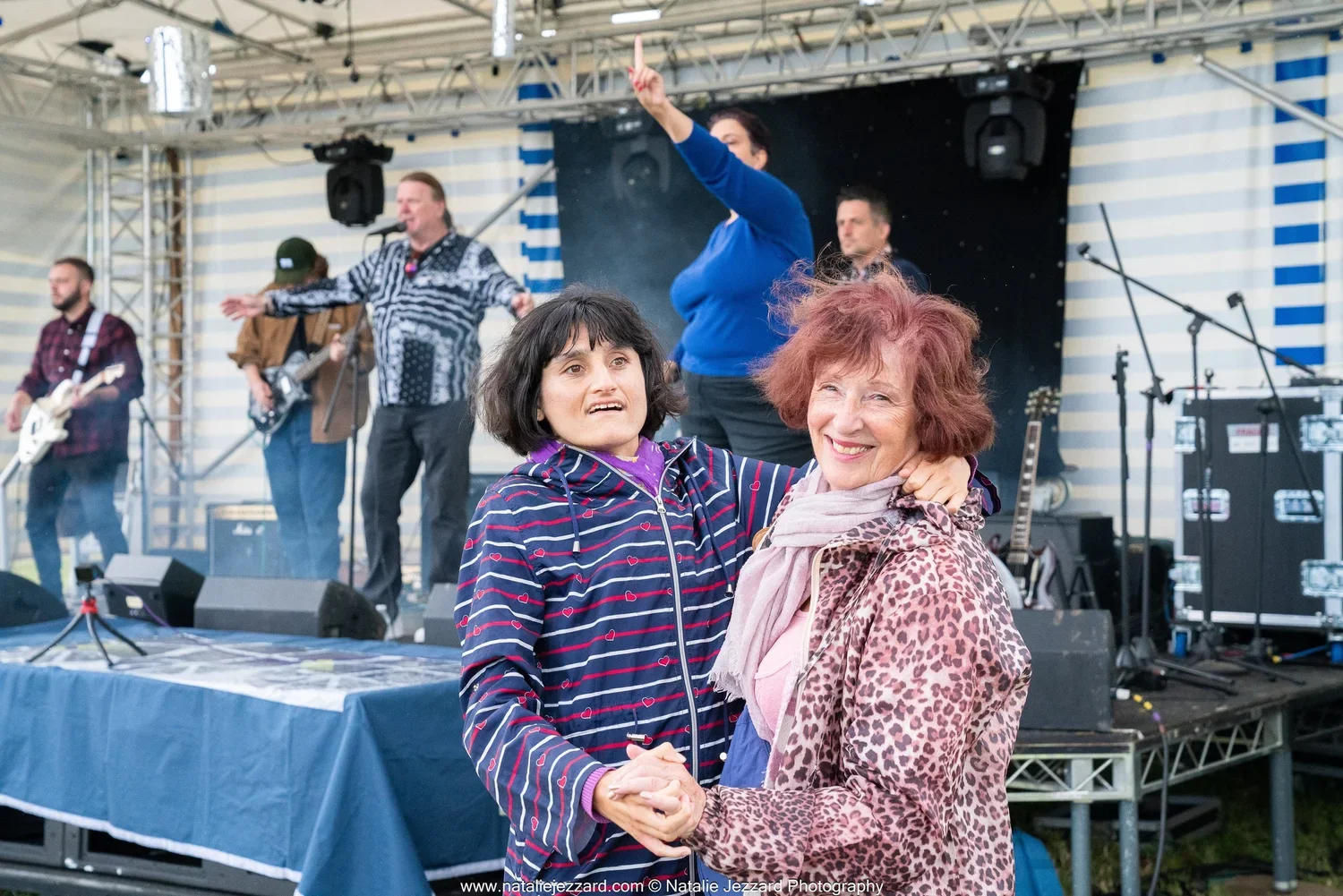 Two women dancing and holding hands in front of a live music band on stage at an outdoor event.