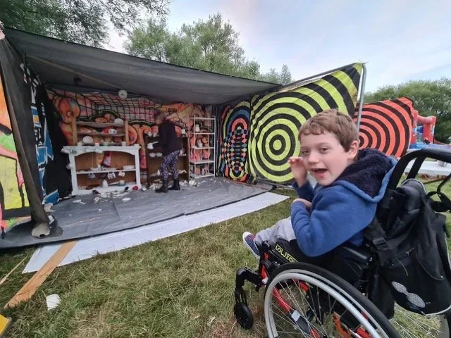 A young boy in a wheelchair smiling outside near a colorful, psychedelic art-covered tent with a person inside and shelves with items.