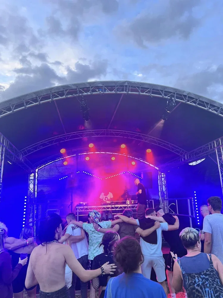 People dancing and enjoying a live outdoor music performance on a stage with colorful lights under a cloudy sky.